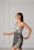 A woman with long dark hair is wearing a stylish, modern dress in soft gray tones. The dress features a unique asymmetrical design with intricate embroidered patterns in darker shades. She is posing gracefully against a minimalist light gray background with panel details.