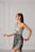 A woman with long dark hair is wearing a stylish, modern dress in soft gray tones. The dress features a unique asymmetrical design with intricate embroidered patterns in darker shades. She is posing gracefully against a minimalist light gray background with panel details.