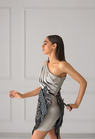 A sleek, elegant woman wearing a flowing, modern dress in soft pastel tones, standing against a minimalist backdrop.