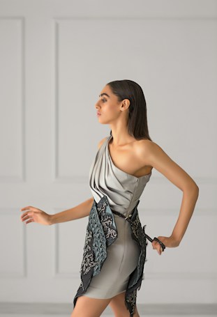 A woman with long dark hair is wearing a stylish, modern dress in soft gray tones. The dress features a unique asymmetrical design with intricate embroidered patterns in darker shades. She is posing gracefully against a minimalist light gray background with panel details.