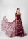 A woman stands elegantly in a studio setting, showcasing a long and flowing maroon gown adorned with small floral embellishments. The dress features a strapless design with intricate detailing on the bodice and a sheer overlay that creates a dramatic train.