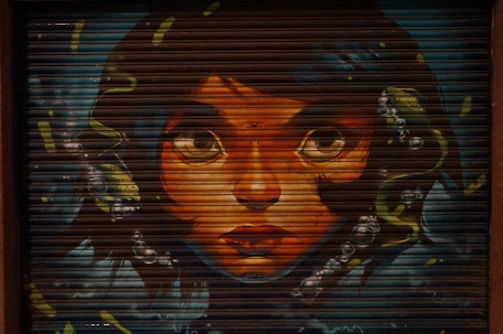 A vivid graffiti mural painted on a metal shutter, depicting a woman's face with large, expressive eyes. The artwork is detailed, with flowing dark hair adorned with bubbles and abstract shapes.