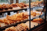 A pastry display case filled with various types of bread and baked goods, including baguettes, rolls, pretzels, and pastries with different shapes and textures. The baked items are arranged in rows on wooden shelves behind a glass front, with a variety of golden brown hues indicating a freshly baked assortment.