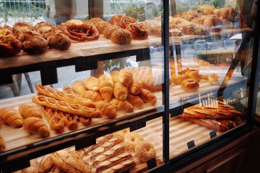 A pastry display case filled with various types of bread and baked goods, including baguettes, rolls, pretzels, and pastries with different shapes and textures. The baked items are arranged in rows on wooden shelves behind a glass front, with a variety of golden brown hues indicating a freshly baked assortment.