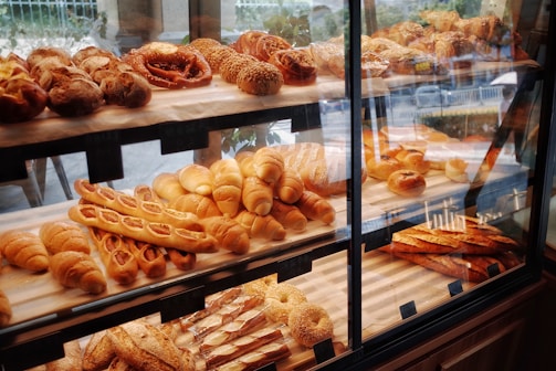 A pastry display case filled with various types of bread and baked goods, including baguettes, rolls, pretzels, and pastries with different shapes and textures. The baked items are arranged in rows on wooden shelves behind a glass front, with a variety of golden brown hues indicating a freshly baked assortment.