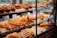A pastry display case filled with various types of bread and baked goods, including baguettes, rolls, pretzels, and pastries with different shapes and textures. The baked items are arranged in rows on wooden shelves behind a glass front, with a variety of golden brown hues indicating a freshly baked assortment.