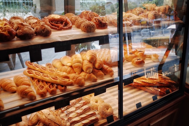 A pastry display case filled with various types of bread and baked goods, including baguettes, rolls, pretzels, and pastries with different shapes and textures. The baked items are arranged in rows on wooden shelves behind a glass front, with a variety of golden brown hues indicating a freshly baked assortment.
