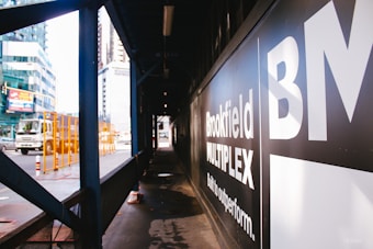 An urban construction area with a sign displaying 'Brookfield Multiplex' on a black background. A truck is visible on the street with orange safety barriers nearby. Skyscrapers and other buildings can be seen in the background.
