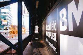 An urban construction area with a sign displaying 'Brookfield Multiplex' on a black background. A truck is visible on the street with orange safety barriers nearby. Skyscrapers and other buildings can be seen in the background.