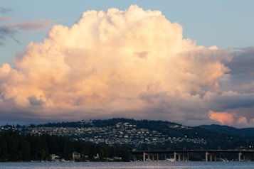 A large, fluffy cloud dominates the sky over a dense hillside neighborhood. The cloud is illuminated with a warm, golden hue, hinting at sunset. Below, a bridge spans across a body of water surrounded by lush trees.