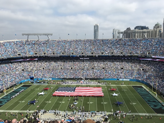 A large football stadium filled with spectators. The field features a giant American flag, surrounded by people. The stands are packed with fans, and there are skyscrapers visible in the city skyline behind the stadium.