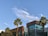 A modern building with large glass windows is framed by several palm trees under a clear blue sky with wispy clouds.