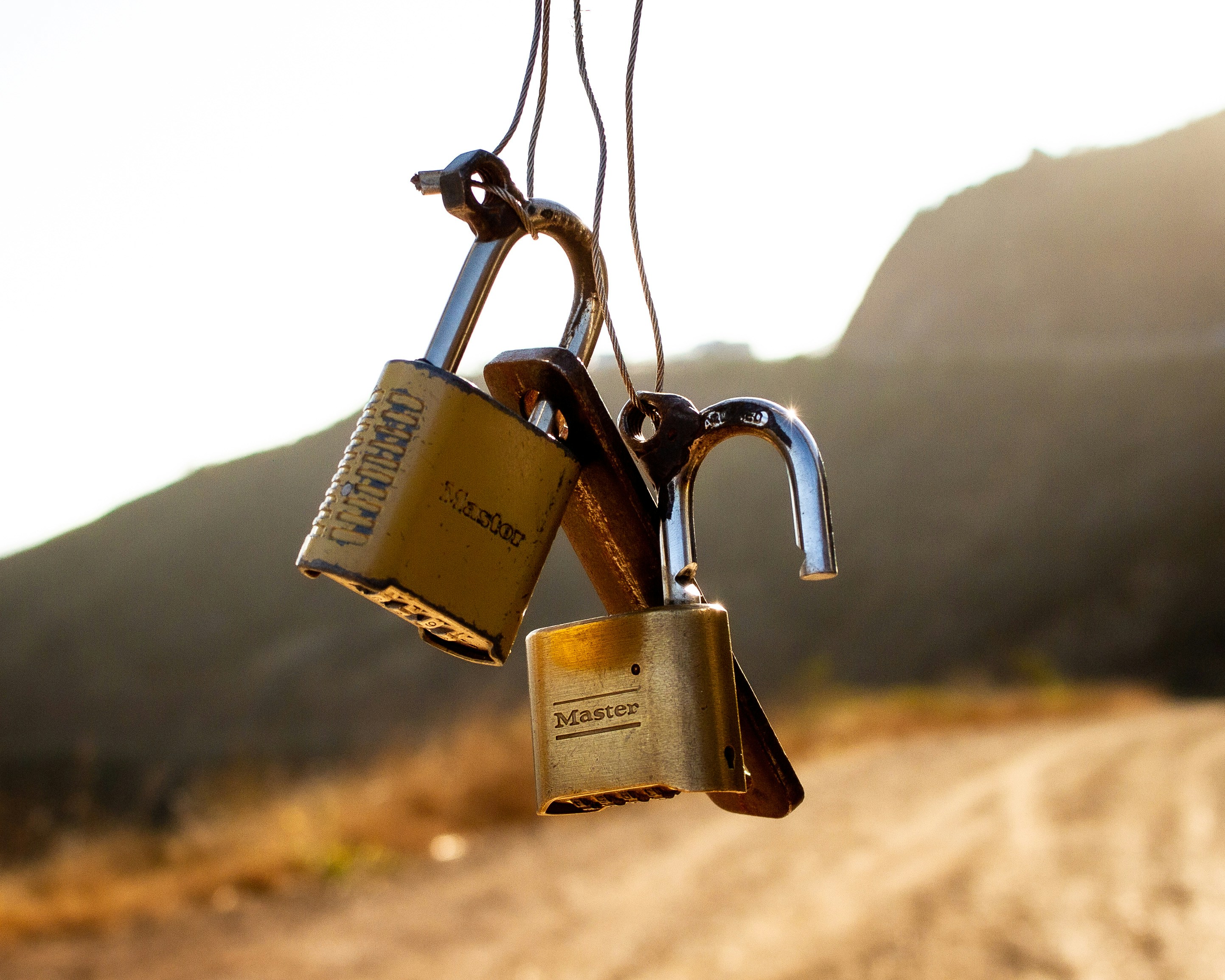 Two brass padlocks hanging from wires against a dry hillside, one of them open