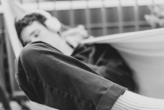 A person reclines in a hammock listening to music through headphones. The image is captured in black and white, highlighting the relaxed posture and casual clothing, such as a pair of dark pants and light socks.