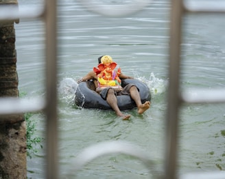 A person is floating on an inner tube in a body of water, holding a colorful statue of Ganesha. The water has gentle ripples, and there are some splashes around the tube. The scene is viewed through a barred structure, adding depth to the perspective.