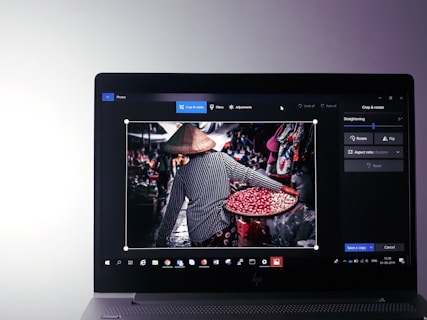 A laptop screen displaying a photo editing software with an image of a person holding a tray filled with red beans in a market setting. The person is wearing a striped shirt and a traditional conical hat, facing away from the camera.