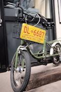A green bicycle is equipped with a metal basket attached to the front, which holds a decorative yellow license plate that reads 'DL 6687 Land of Enchantment 80 New Mexico'. The bike is parked on a wooden block in front of a building with black-framed windows.