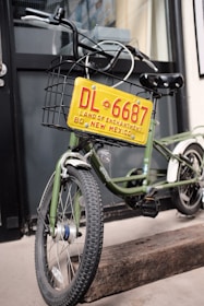 A green bicycle is equipped with a metal basket attached to the front, which holds a decorative yellow license plate that reads 'DL 6687 Land of Enchantment 80 New Mexico'. The bike is parked on a wooden block in front of a building with black-framed windows.