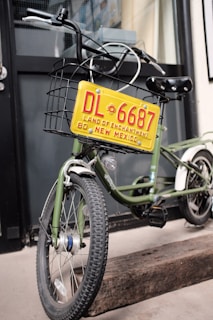 A green bicycle is equipped with a metal basket attached to the front, which holds a decorative yellow license plate that reads 'DL 6687 Land of Enchantment 80 New Mexico'. The bike is parked on a wooden block in front of a building with black-framed windows.
