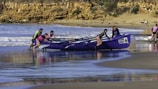 A team of people is pushing a boat labeled 'TEAM NAVY' into the water from a sandy beach. The ocean waves are gently crashing in the background, and the scene is set against a backdrop of rocky cliffs and scattered greenery. The team members are wearing brightly colored outfits, including life vests and caps, suggesting that they are preparing for a rowing event or training exercise.