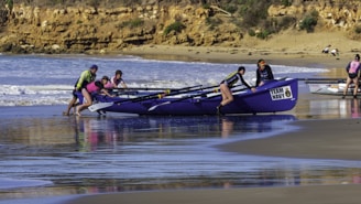 A team of people is pushing a boat labeled 'TEAM NAVY' into the water from a sandy beach. The ocean waves are gently crashing in the background, and the scene is set against a backdrop of rocky cliffs and scattered greenery. The team members are wearing brightly colored outfits, including life vests and caps, suggesting that they are preparing for a rowing event or training exercise.