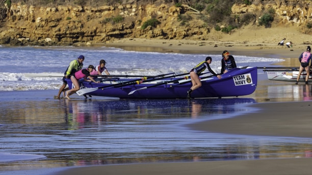 A team of people is pushing a boat labeled 'TEAM NAVY' into the water from a sandy beach. The ocean waves are gently crashing in the background, and the scene is set against a backdrop of rocky cliffs and scattered greenery. The team members are wearing brightly colored outfits, including life vests and caps, suggesting that they are preparing for a rowing event or training exercise.