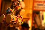 A collection of intricately crafted miniature drums hangs closely together, showcasing colorful designs and patterns. The ambient lighting casts a warm glow that highlights the textures and materials used in their construction, with a defocused background indicating an indoor setting possibly in a market or shop.