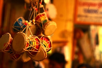 A collection of intricately crafted miniature drums hangs closely together, showcasing colorful designs and patterns. The ambient lighting casts a warm glow that highlights the textures and materials used in their construction, with a defocused background indicating an indoor setting possibly in a market or shop.