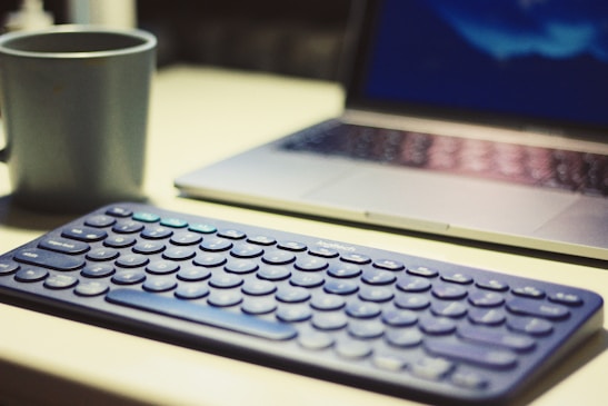 A computer setup featuring a wireless keyboard, a laptop, and a coffee mug on a desk. The keyboard is prominently placed in the foreground, while the laptop screen is slightly blurred in the background. The mug is positioned to the left of the keyboard.