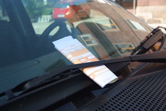 A parking ticket is tucked under the windshield wiper of a car. The background shows a red car parked in an outdoor setting.