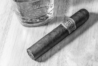 A glass tumbler with embossed details next to a dark cigar on a wooden surface. The cigar has a label wrapped around it displaying text. The lighting highlights the textures of the wood and the items.