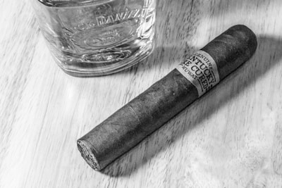 A glass tumbler with embossed details next to a dark cigar on a wooden surface. The cigar has a label wrapped around it displaying text. The lighting highlights the textures of the wood and the items.
