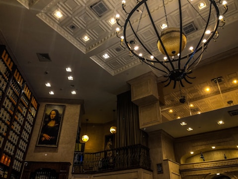 An elegant interior with a high ceiling adorned with an intricate metal chandelier. The walls are lined with shelves filled with wine bottles. A large framed portrait resembling the Mona Lisa is prominently displayed on one wall, adding a classic touch to the decor. Warm lighting creates a cozy and sophisticated atmosphere.