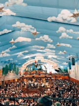 A large hall filled with a festive crowd under a tent ceiling painted with blue sky and clouds, adorned with stars. Central to the scene is a wooden structure decorated with greenery and flags, with people standing and observing the activities below. The walls also feature murals of landscapes and buildings.
