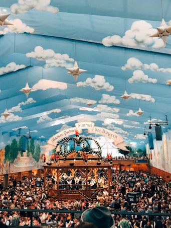 A large hall filled with a festive crowd under a tent ceiling painted with blue sky and clouds, adorned with stars. Central to the scene is a wooden structure decorated with greenery and flags, with people standing and observing the activities below. The walls also feature murals of landscapes and buildings.