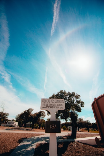 A signpost in a rural landscape under a bright blue sky with scattered clouds and contrails. The sign contains text in Portuguese and stands on a paved path surrounded by sparse trees. There's a structure partially visible on the right side.