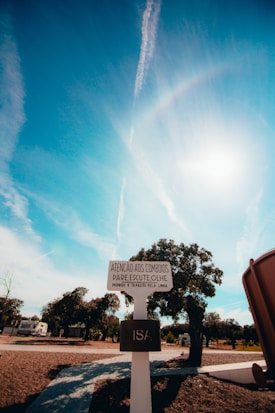 A signpost in a rural landscape under a bright blue sky with scattered clouds and contrails. The sign contains text in Portuguese and stands on a paved path surrounded by sparse trees. There's a structure partially visible on the right side.