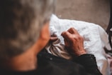 An elderly person is sewing or embroidering on a piece of white fabric, with detailed focus on their hands. The lighting is warm and soft, highlighting the texture of the skin and fabric.