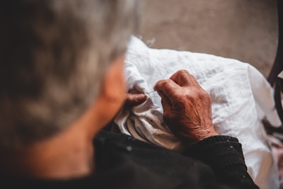 An elderly person is sewing or embroidering on a piece of white fabric, with detailed focus on their hands. The lighting is warm and soft, highlighting the texture of the skin and fabric.