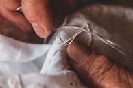 A close-up of hands engaged in sewing, with fingers carefully guiding a needle and thread through white fabric. The hands, slightly aged, convey a sense of dexterity and focus.