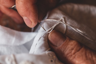 A close-up of hands engaged in sewing, with fingers carefully guiding a needle and thread through white fabric. The hands, slightly aged, convey a sense of dexterity and focus.