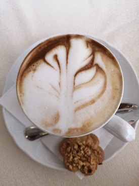 A cup of cappuccino with intricate latte art in the shape of a leaf on top. The cup is placed on a white saucer with a small spoon to the side. A cookie with nuts is on the saucer beside the cup, resting on a folded napkin.