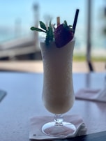 A tall glass filled with a creamy white frozen drink, garnished with a sprig of mint and a whole strawberry. Two straws are inserted into the drink. The background features a blurred outdoor scene, likely by a beach or poolside.