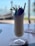 A tall glass filled with a creamy white frozen drink, garnished with a sprig of mint and a whole strawberry. Two straws are inserted into the drink. The background features a blurred outdoor scene, likely by a beach or poolside.