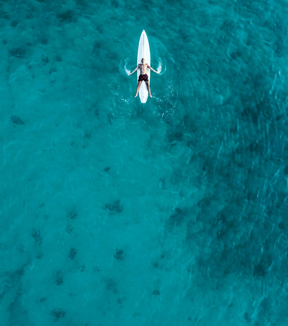 man wears board shorts lying on white surfboard at the beach