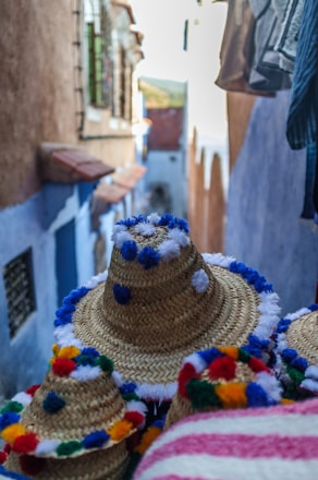 Colorful woven straw hats with multicolored pom-poms are in the foreground. The background shows a narrow street with vibrant blue and white walls, typical of a traditional Mediterranean or North African village.