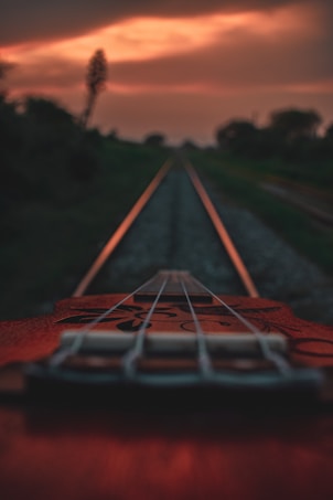 A guitar is positioned along a set of railway tracks, creating a creative blend of musical elements and industrial lines. The focus is on the guitar strings with the tracks leading towards a glowing orange sunset sky.