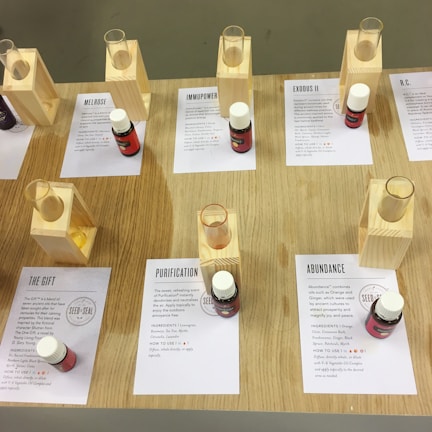 A display of essential oils in small bottles with accompanying information cards and wooden stands. The wooden table supports multiple sets of these items, with the oils labeled with names like 'Melrose', 'ImmuPower', 'Exodus II', 'Purification', 'The Gift', and 'Abundance'. Each oil bottle is placed next to a card outlining its ingredients and uses.