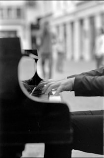 A black and white photograph captures a person playing a grand piano. The focus is on the hands and keys, with a blurred background revealing an urban setting.