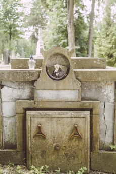 An old, weathered tombstone with a photograph of a man in formal attire is embedded in the center. The tombstone features inscriptions and has signs of aging, like moss and cracks. There is a small, decorative vase or candle holder placed on the stone. The background shows a lush, green cemetery with several trees and other gravestones.